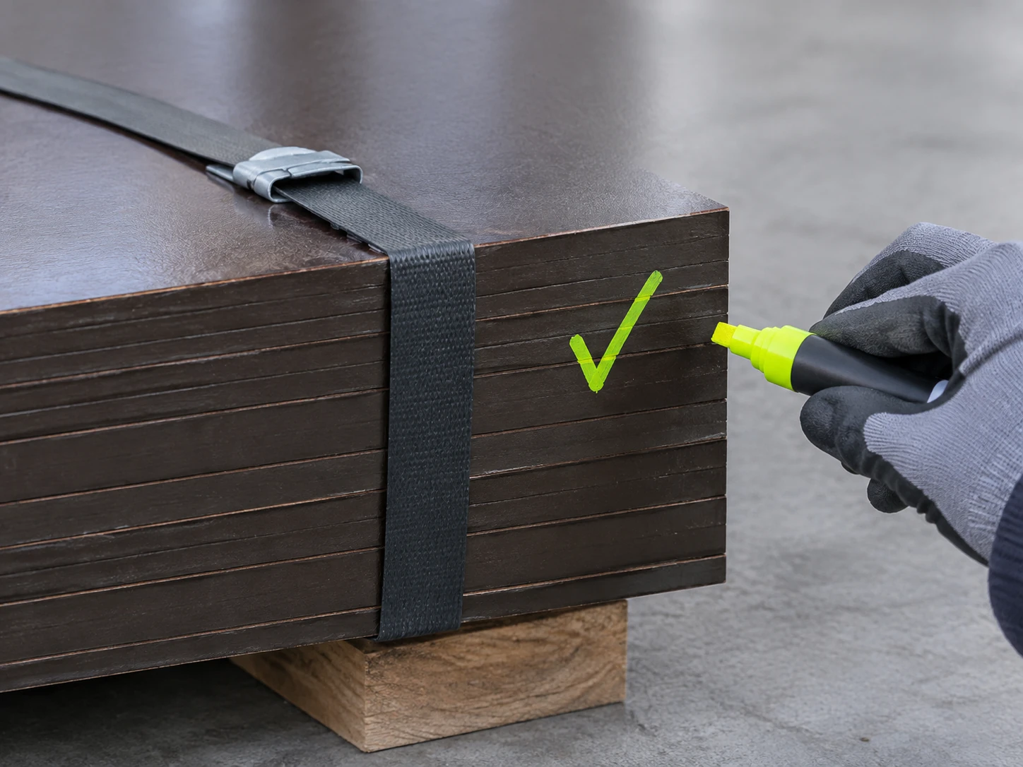 Gloved hand with a lime paint marker beside a tick drawn on the dark painted edge of a strapped plywood bundle